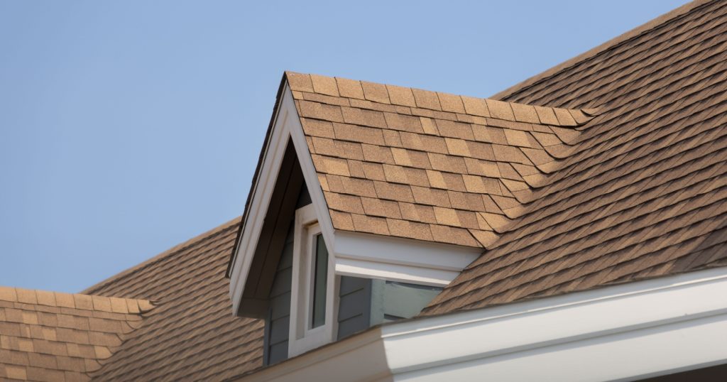 Close-up of a house roof with new brown asphalt shingles and a gable dormer against a clear blue sky