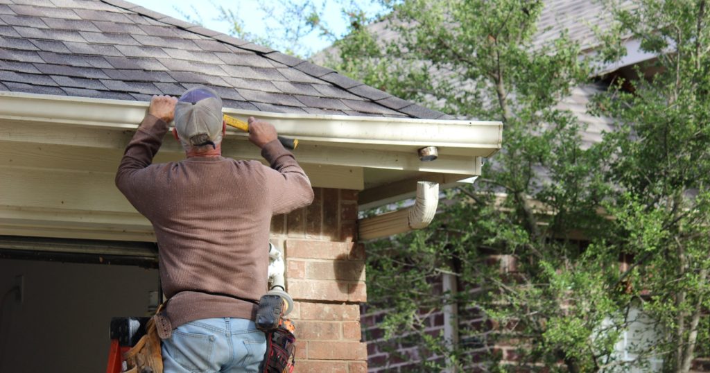 A man standing on a ladder repairing his roof and rain gutters.